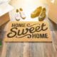 Welcome mat that reads "Home Sweet Home" surrounded by moving boxes and two pairs of shoes