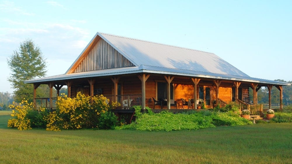 Una cabaña de troncos con techo de metal y un porche envolvente rodeado de arbustos y pastos