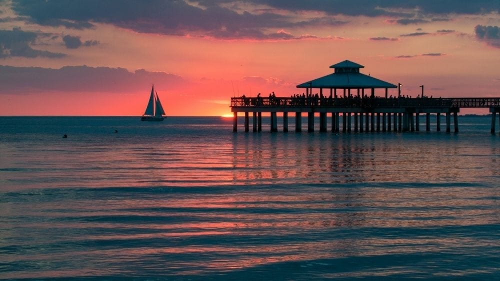 View from the coast of the sun on the horizon, lighting up a pier and a sailboat in the distance.