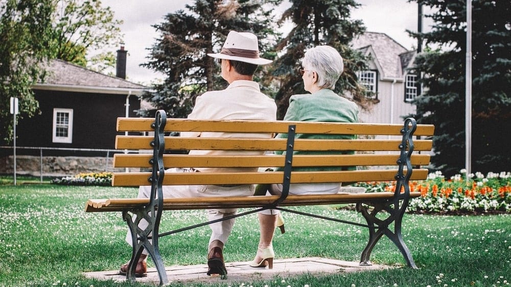 Older couple sitting on bench outside of houses.