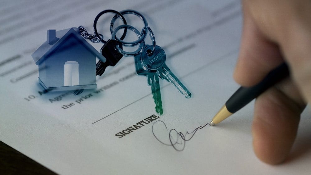 Un llavero de la casa con dos llaves se encuentra encima de un contrato con una mano firmando el documento