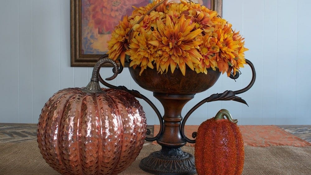 Two decorative pumpkins beside an ornate, rustic vase filled with yellow flowers.