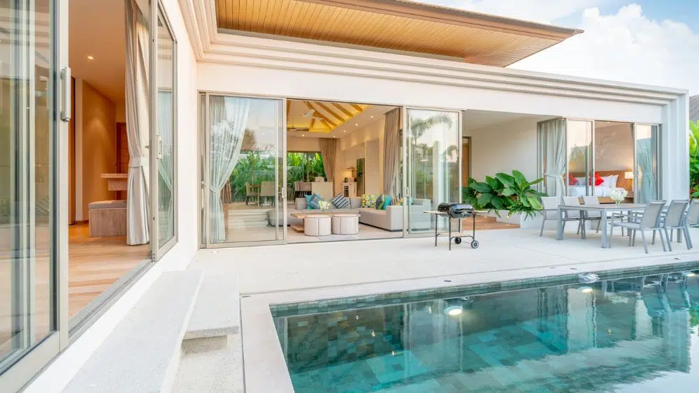 Pool house in a villa style with an open breezeway for a living room and a dining space. Other parts of the house, including a bedroom, are open to the pool via modern sliding glass doors.