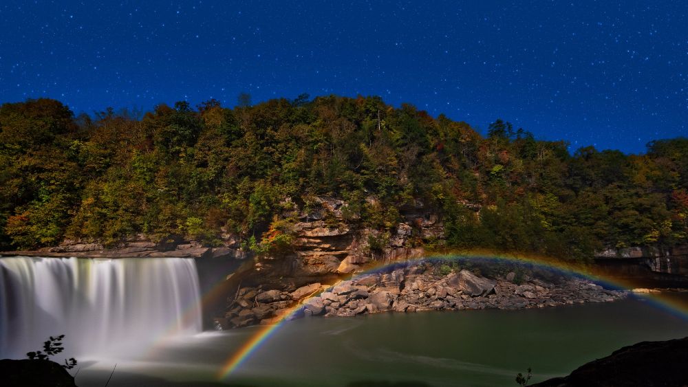 A moonbow over a river with a waterfall. The river is bordered by a forest.