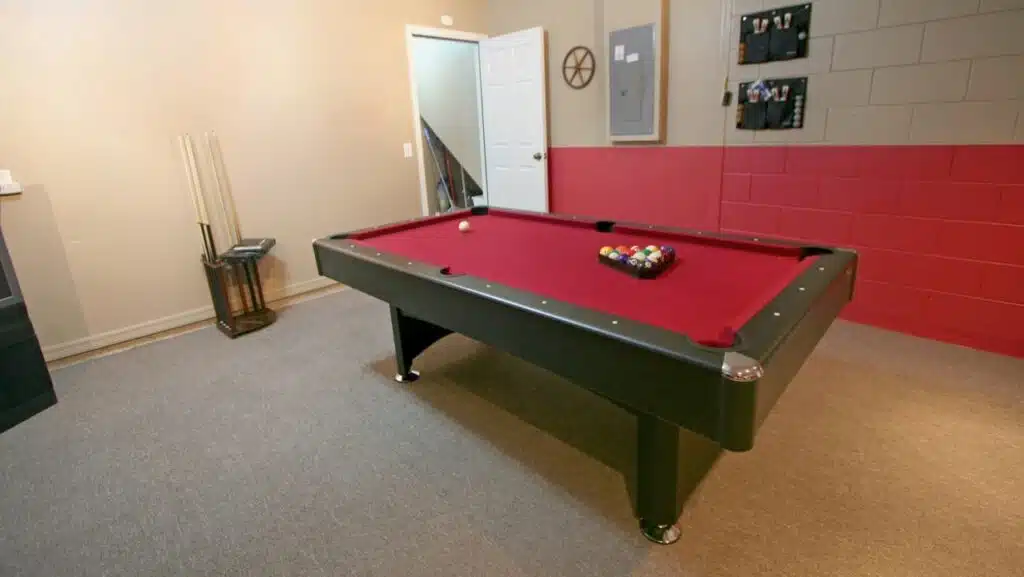 A converted home garage with a red felt covered pool table, brown carpet, and one wall painted red on the bottom and cream on the top.